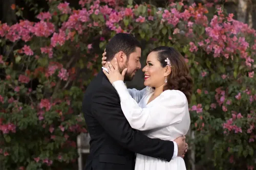 Novios besándose frente a flores rosadas. Imagen romántica con un fondo floral vibrante por David Sánchez.