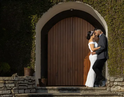  Novios abrazándose frente a una gran puerta de madera rodeada de vegetación. Fotografía rústica y elegante de boda por David Sánchez.